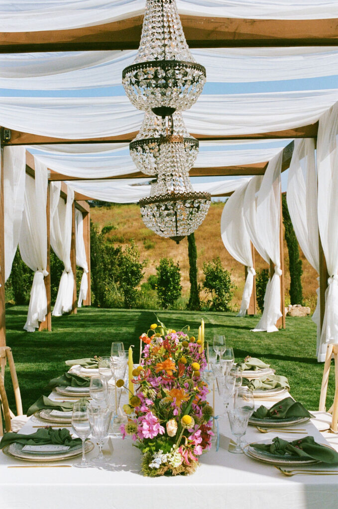 Jeune Perche wedding venue showcasing an elegant table setting with green napkins, crystal glassware, and vibrant floral arrangements. The table is set under a canopy of flowing white drapery, creating a sophisticated and romantic atmosphere against the backdrop of the scenic landscape