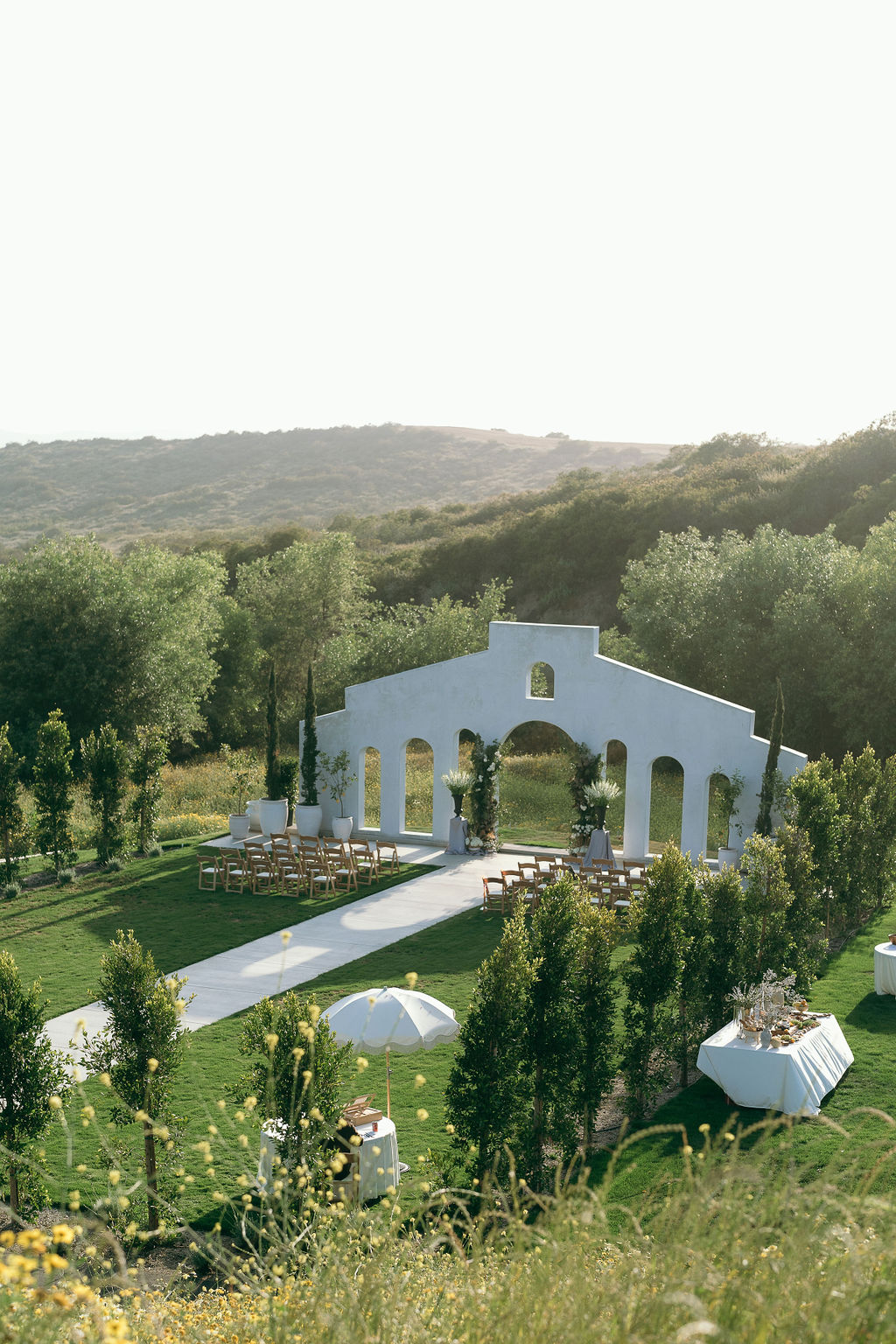 Jeune Perche wedding venue featuring a stunning white concrete archway reminiscent of Mediterranean architecture. The archway is set against a backdrop of lush greenery and rolling hills, with neatly arranged chairs and tables ready for a beautiful outdoor ceremony