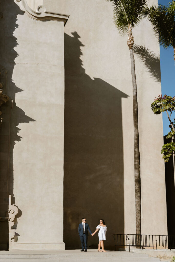 San Diego wedding photographer captures a couple holding hands at Balboa Park. The couple stands against the backdrop of a tall, shadowed wall and palm trees, creating a striking and artistic composition in front of the Museum of Man.