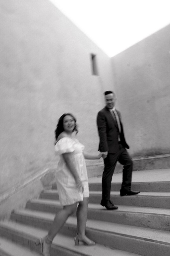 San Diego wedding photographer captures a candid moment of a couple walking up stairs at Balboa Park. The bride wears a white dress, and the groom is in a suit, both smiling and holding hands in a dynamic, black-and-white shot.