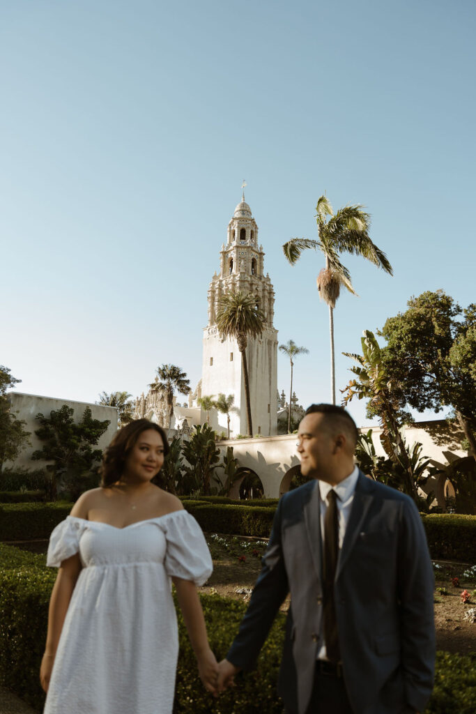 San Diego wedding photographer captures a couple holding hands in front of the iconic California Tower at Balboa Park. The bride wears a white dress, and the groom is in a suit, with the beautiful architecture and lush greenery of the park in the background.