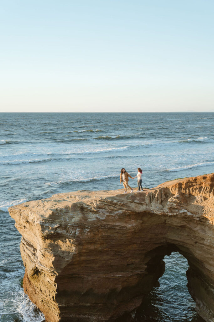 San Diego wedding photographer captures a couple at Sunset Cliffs, standing on a rocky outcrop above the ocean. The couple holds hands, enjoying the breathtaking coastal views and the expansive ocean horizon."