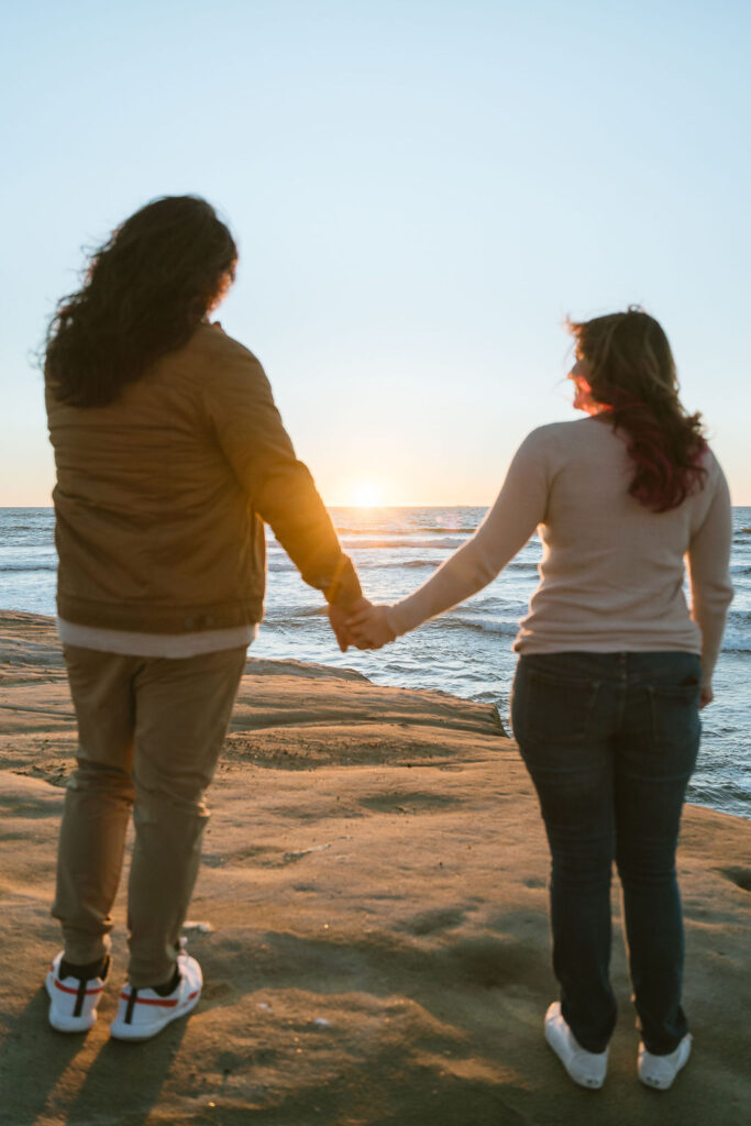 San Diego wedding photographer captures a couple holding hands while watching the sunset at Sunset Cliffs. The romantic moment highlights the couple's silhouette against the setting sun and the serene ocean waves