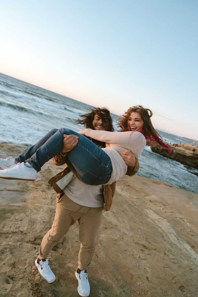 San Diego wedding photographer captures a joyful moment at Sunset Cliffs with a couple laughing as the groom playfully lifts the bride. The scene is set against the backdrop of the ocean and a beautiful sunset.