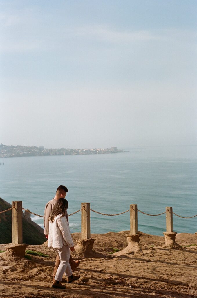 San Diego wedding photographer captures a couple walking along the cliffs at Torrey Pines Gliderport. The couple strolls hand-in-hand, enjoying the stunning coastal view of the ocean and the distant cityscape