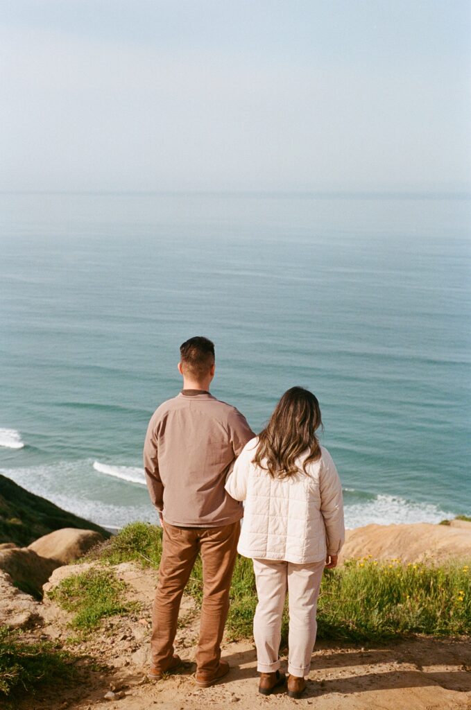 San Diego wedding photographer captures a couple admiring the ocean view at Torrey Pines Gliderport. The couple stands side by side on the cliff's edge, gazing out at the calm sea and the clear blue sky