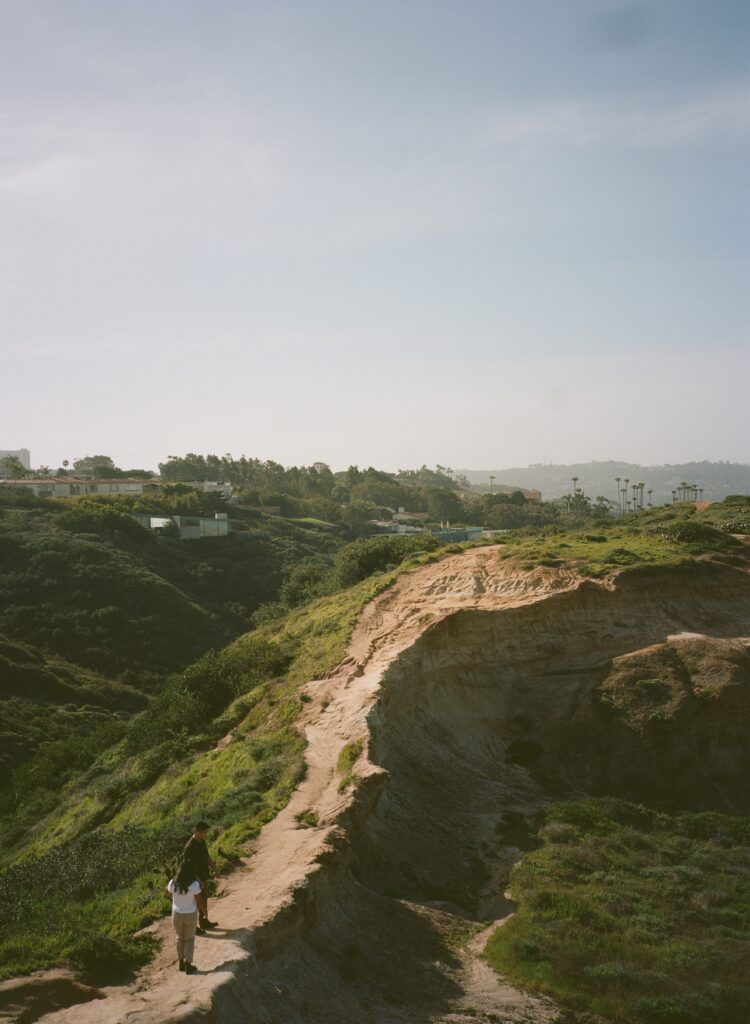 San Diego wedding photographer captures a couple walking along the rugged cliffs at Torrey Pines Gliderport. The couple is seen from a distance, highlighting the dramatic landscape and the beautiful coastal scenery