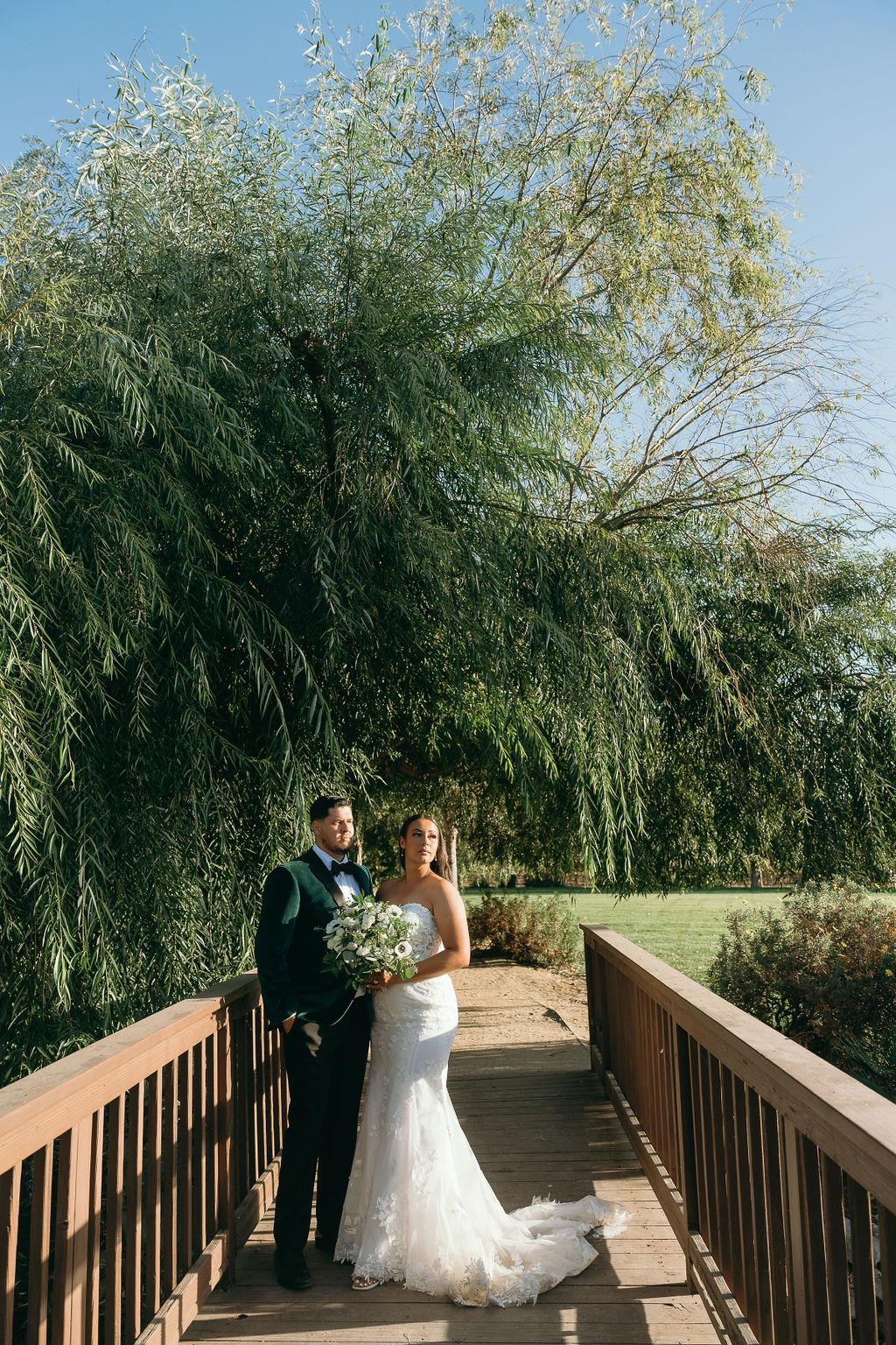 Bride and groom pose under lush willow trees on a wooden bridge at Wolfe Heights Wedding.