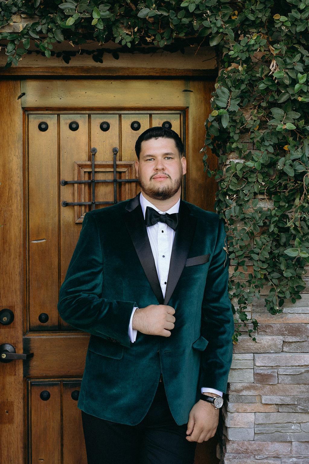 Groom in a green velvet tuxedo standing confidently against a rustic wooden door at Wolfe Heights Wedding