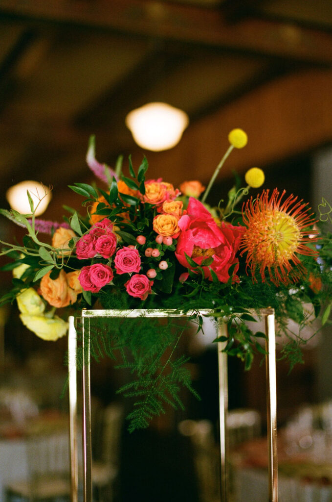 Vibrant wedding centerpiece with pink roses, orange ranunculus, and pincushion protea on a gold stand. A bold floral arrangement at a Northern California wedding reception.