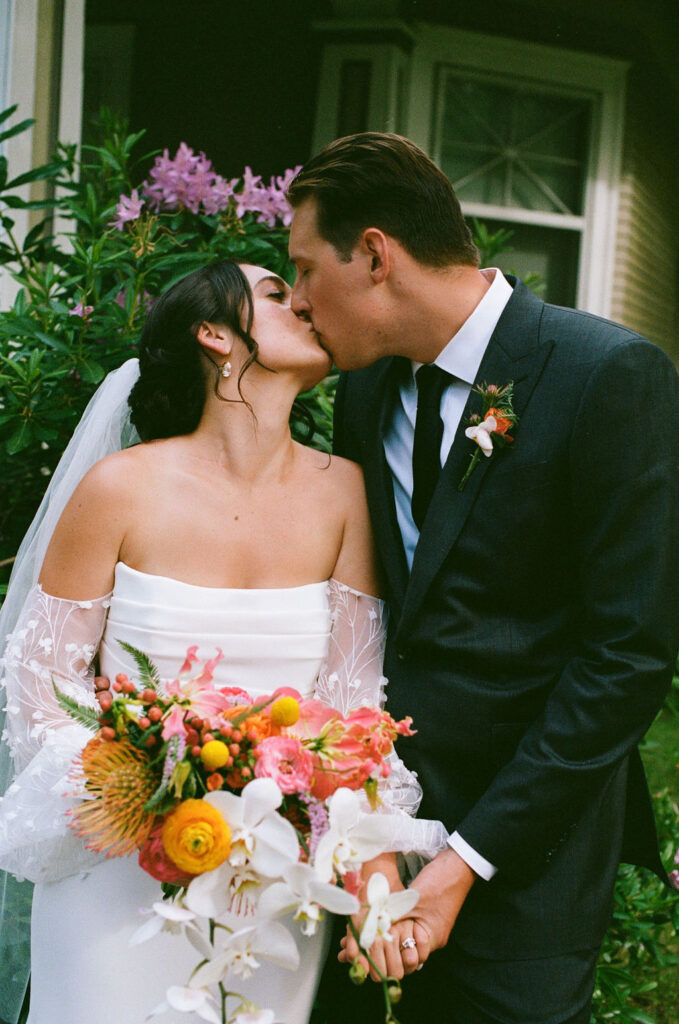 Bride and groom share a kiss in front of blooming flowers, with a vibrant bouquet in hand. Captured on film for a timeless and romantic wedding portrait in Northern California.