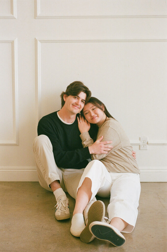 Couple sitting against a white panel wall, captured on 35mm film during a studio engagement session.