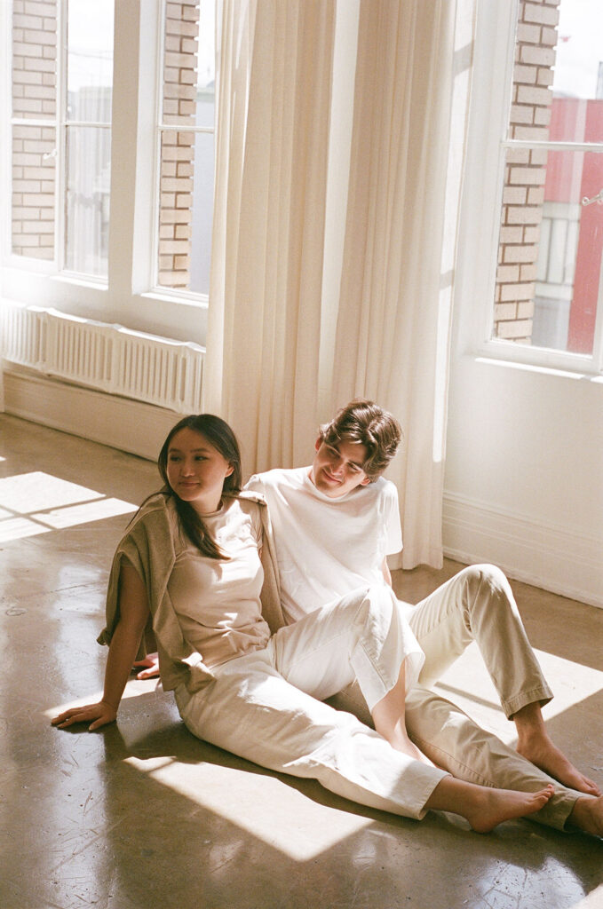 Couple sitting on the floor in a sunlit corner, embracing in soft light during a 35mm film studio engagement session.