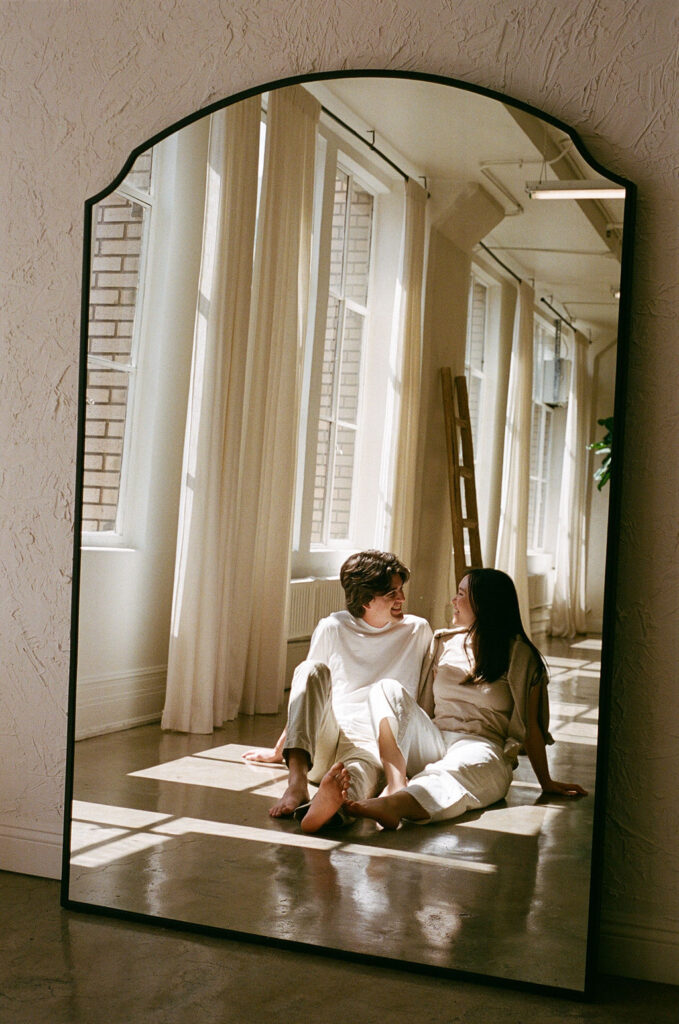 Reflection of a couple sitting together, framed in a large mirror with natural light, captured on 35mm film.