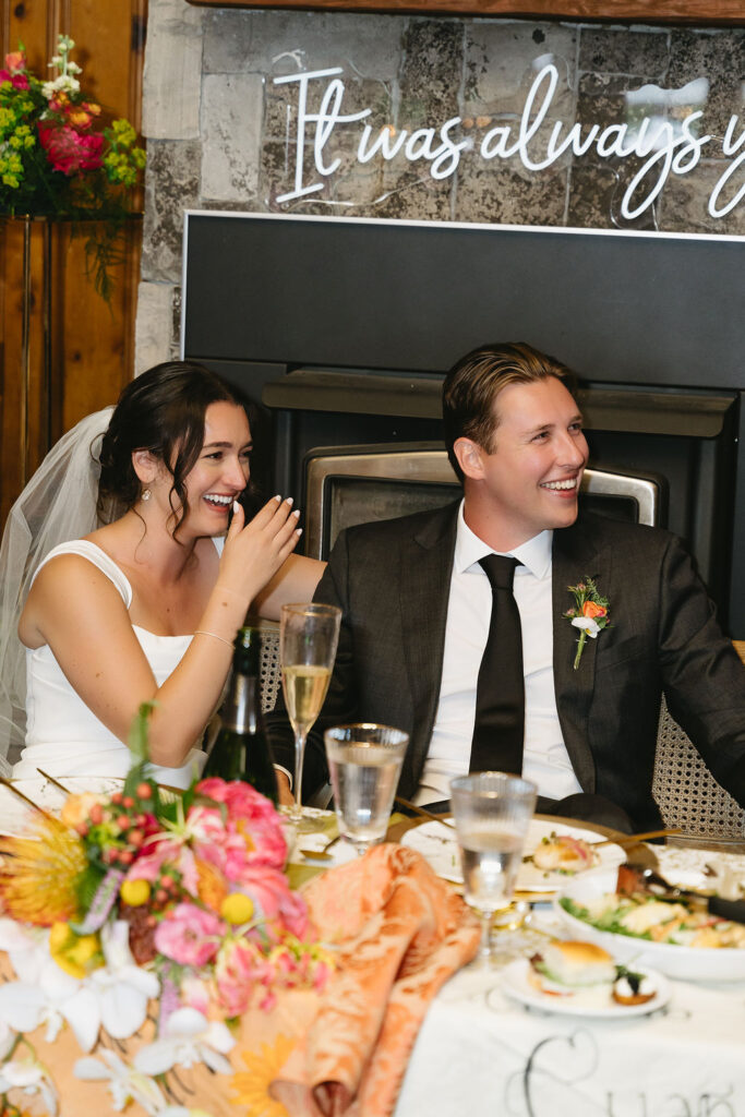 Bride and groom share a laugh during dinner, seated in front of a neon sign reading “It was always you.”