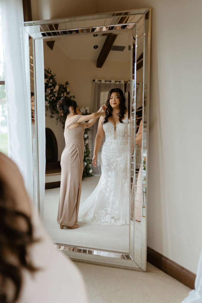 Bride in the mirror as bridesmaid fixes her dress as she gets ready by a window