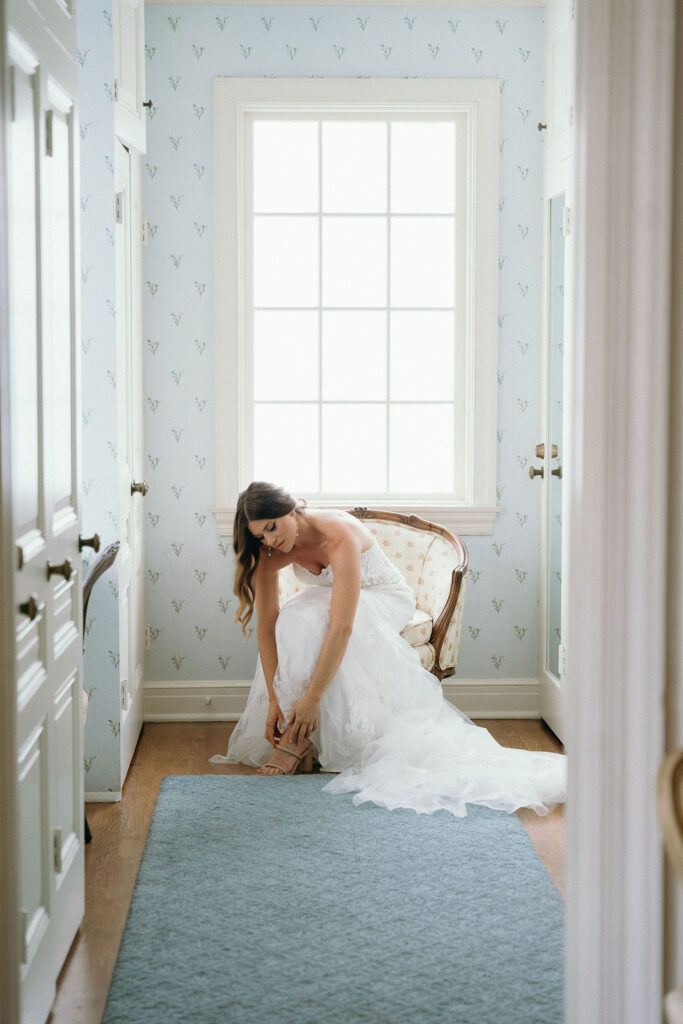 Bride getting ready in a hallway in front of a window putting her heals on