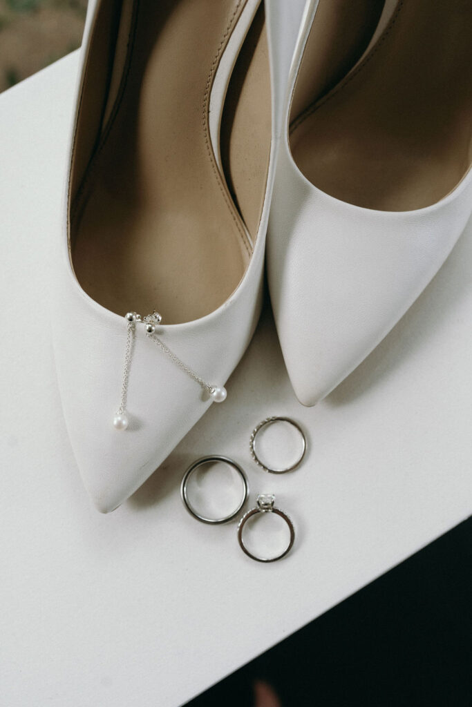 White bridal heels adorned with pearl earrings and wedding bands flatlay during getting ready.