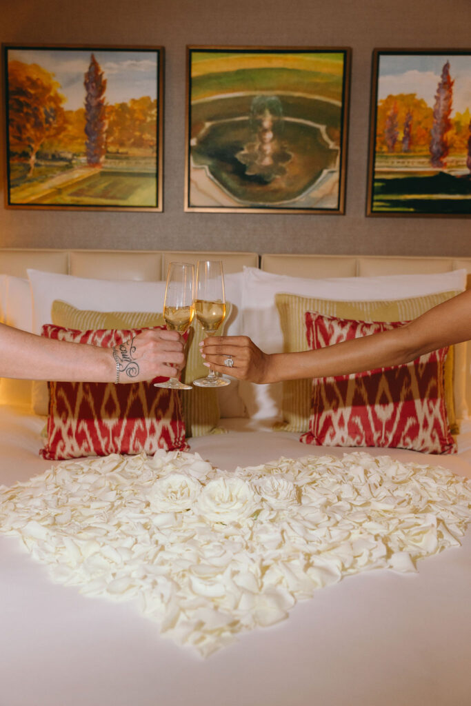 wo hands clinking champagne glasses over a heart-shaped bed of white rose petal