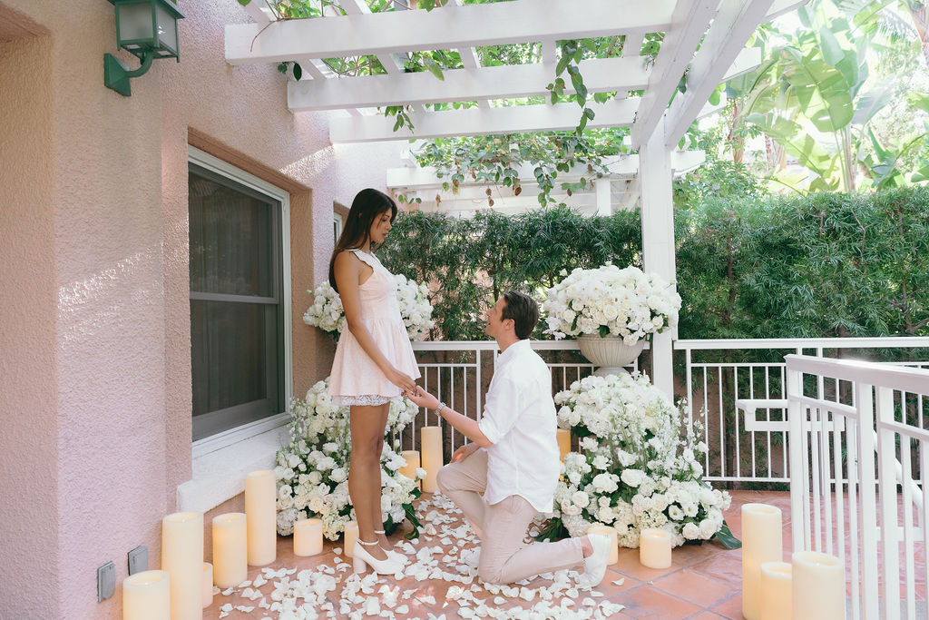 Romantic proposal moment with a groom-to-be kneeling among white floral arrangements in Los Angeles.