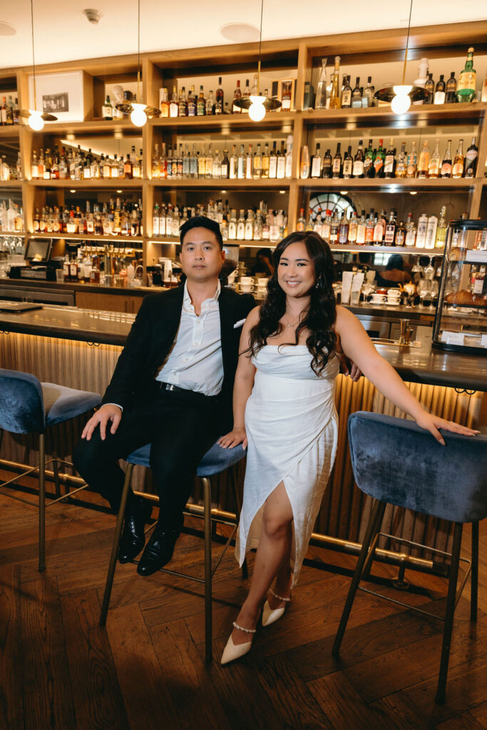 ViLan and Jason sit at a sleek bar with a wall of liquor bottles behind them, capturing a moody engagement vib