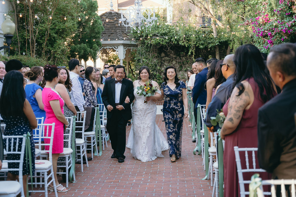 Bride walks down the aisle with her parents at Vizcaya Sacramento, surrounded by guests and lush greener