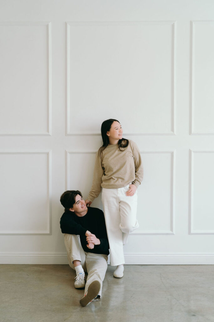 Woman leans against the wall as her partner sits below, both relaxed in a modern engagement session.