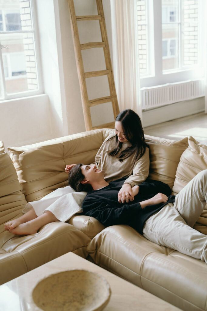 Couple lounging on a tan leather couch in a softly lit studio engagement session.