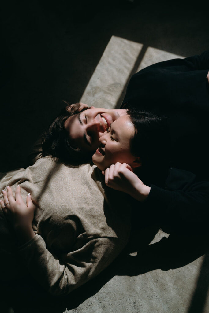 Couple lying on the floor in dramatic direct light, captured in a modern studio engagement session.