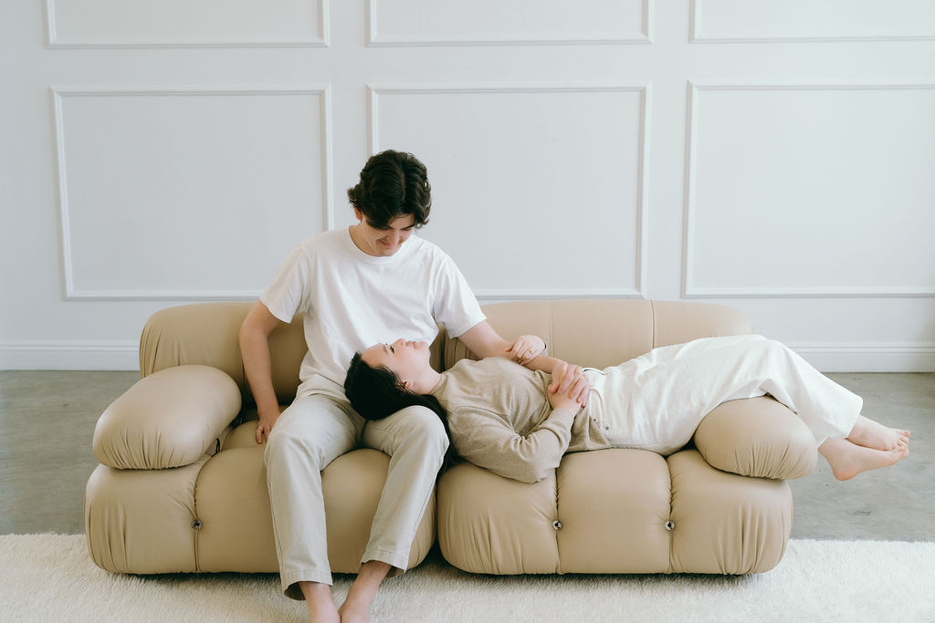 Couple lounging on a tan leather couch in a softly lit studio engagement session.