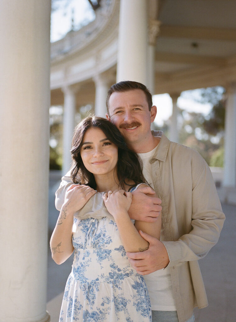 Smiling couple posing together in soft sunlight under white columns at Balboa Park